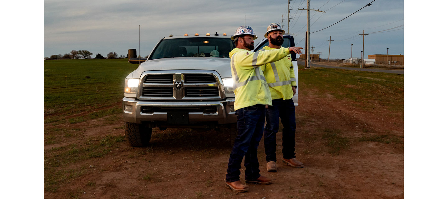 Dos trabajadores de servicios públicos, Kevin Walker y Dylan Edgmon, con camisetas de seguridad amarillas y cascos, están de pie junto a un camión blanco al atardecer; uno de ellos sostiene un portapapeles y señala hacia la distancia.